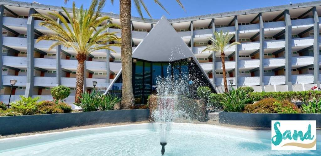 Resort entrance with fountain, triangular glass atrium, palm trees and white multi-storey facade; 'Sand' logo