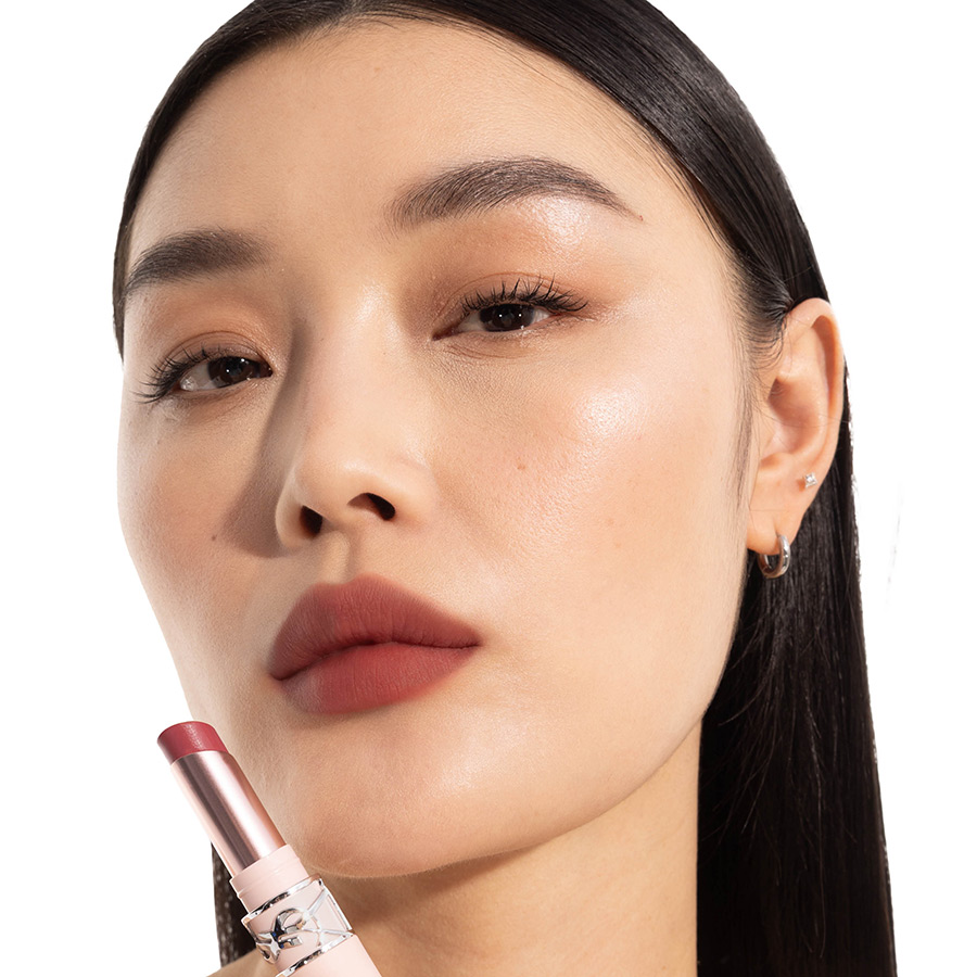 Primo piano di una donna asiatica che mostra un rossetto opaco color legno di rosa, con in mano il rossetto YSL coordinato.