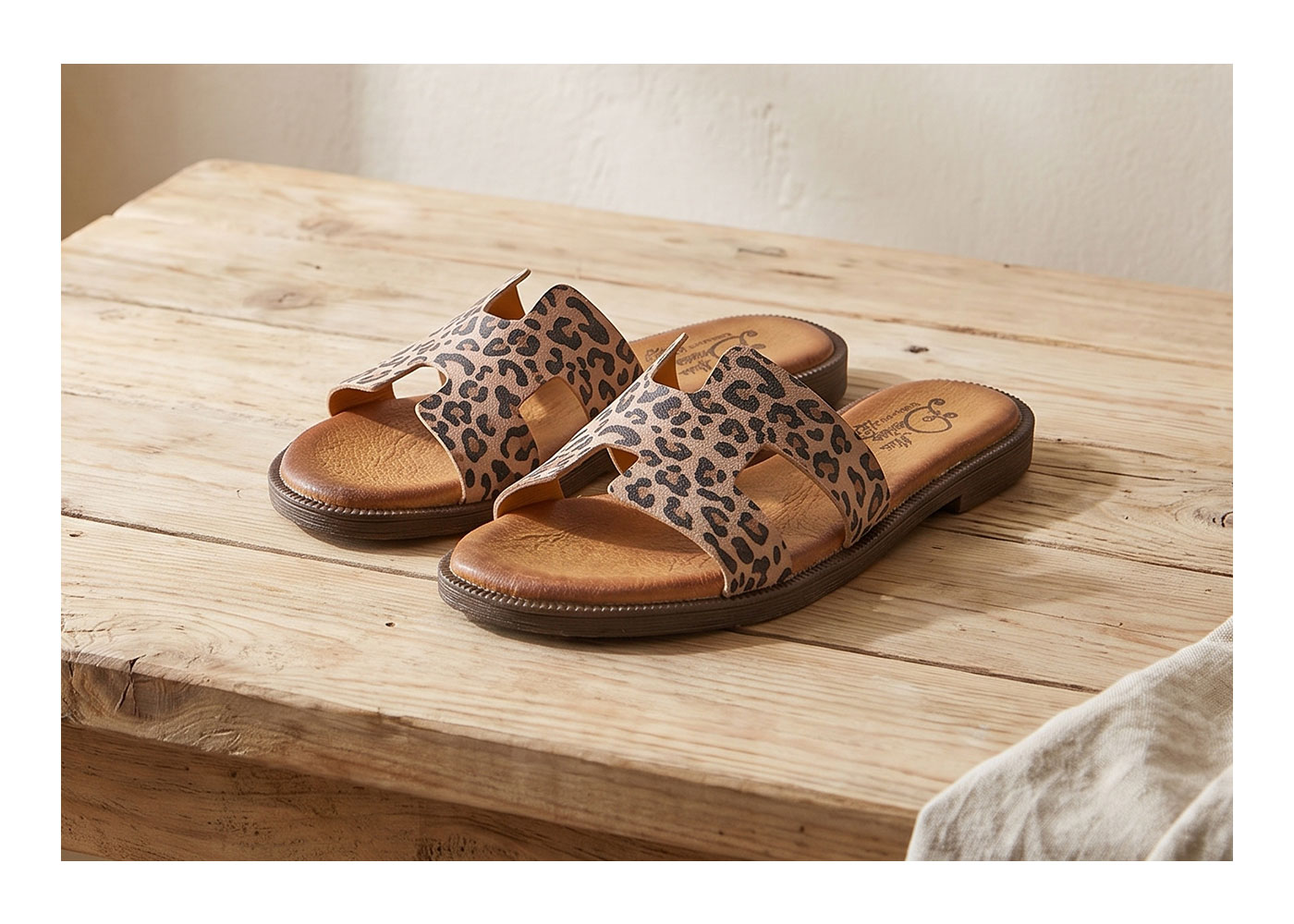 Pair of women's leopard-print leather slide sandals with cushioned tan footbed on rustic wooden table