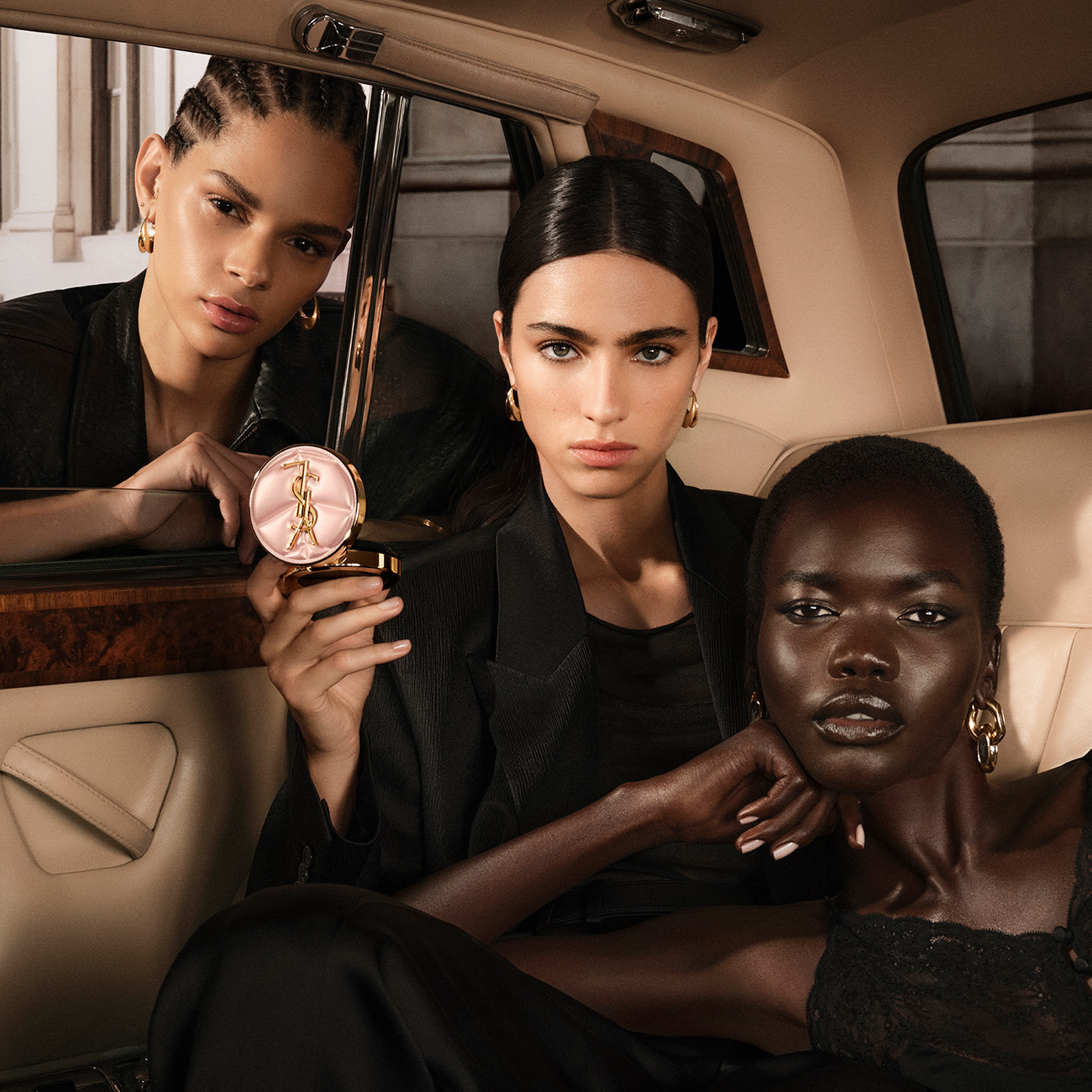 Tre donne eleganti posano all'interno di un'auto d'epoca di lusso, e una di loro tiene in mano un astuccio cushion rosa trapuntato YSL.