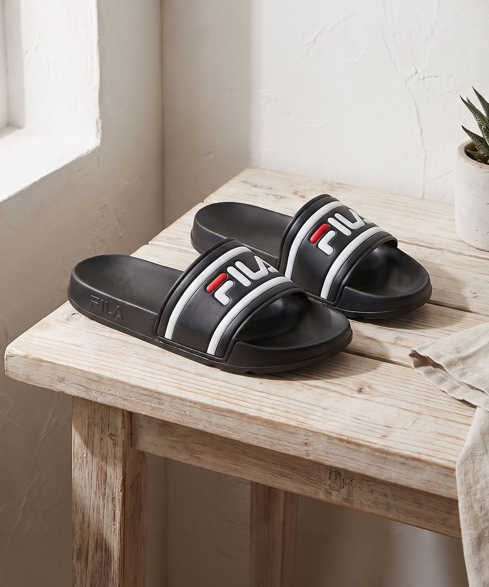 Black FILA slide sandals with white and red logo on a wooden bench by a sunlit window and small potted succulent