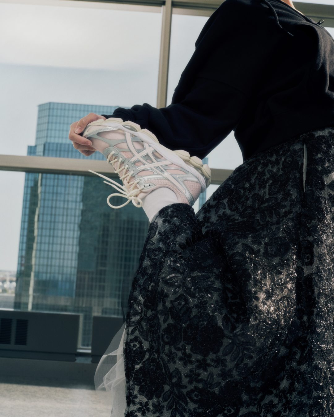 Person in black floral skirt and hoodie holding up white pastel trainers and socks by a floor-to-ceiling city high-rise window