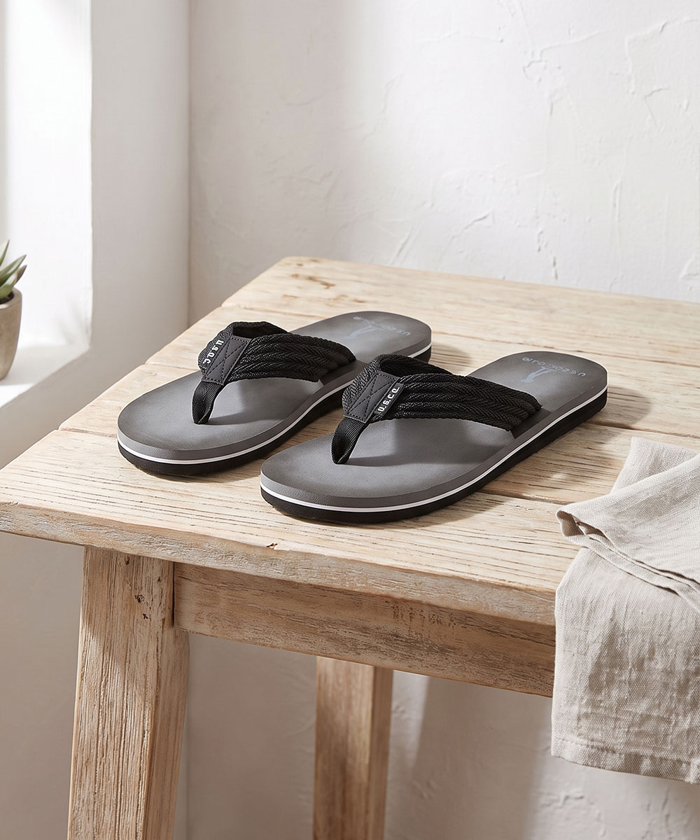 Pair of men's grey foam flip-flops with black braided straps on a rustic wooden bench beside a folded linen cloth by a sunlit window