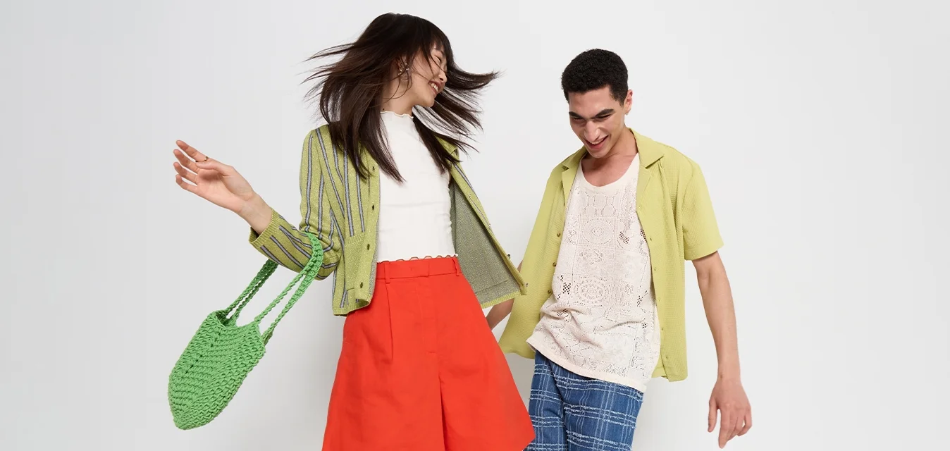 Due modelli posano in vivaci outfit estivi: la donna indossa un cardigan verde a righe, una gonna rossa e una borsa intrecciata verde; l'uomo indossa una canotta beige e pantaloni corti a quadretti blu.