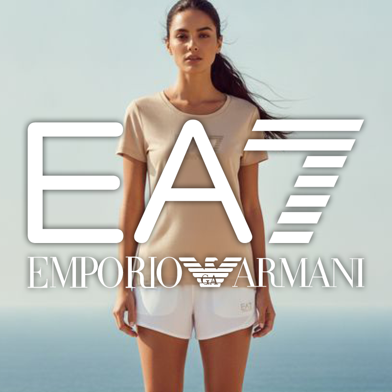 Woman wearing beige T‑shirt and white shorts standing by the sea with a large white EA7 Emporio Armani logo overlaid