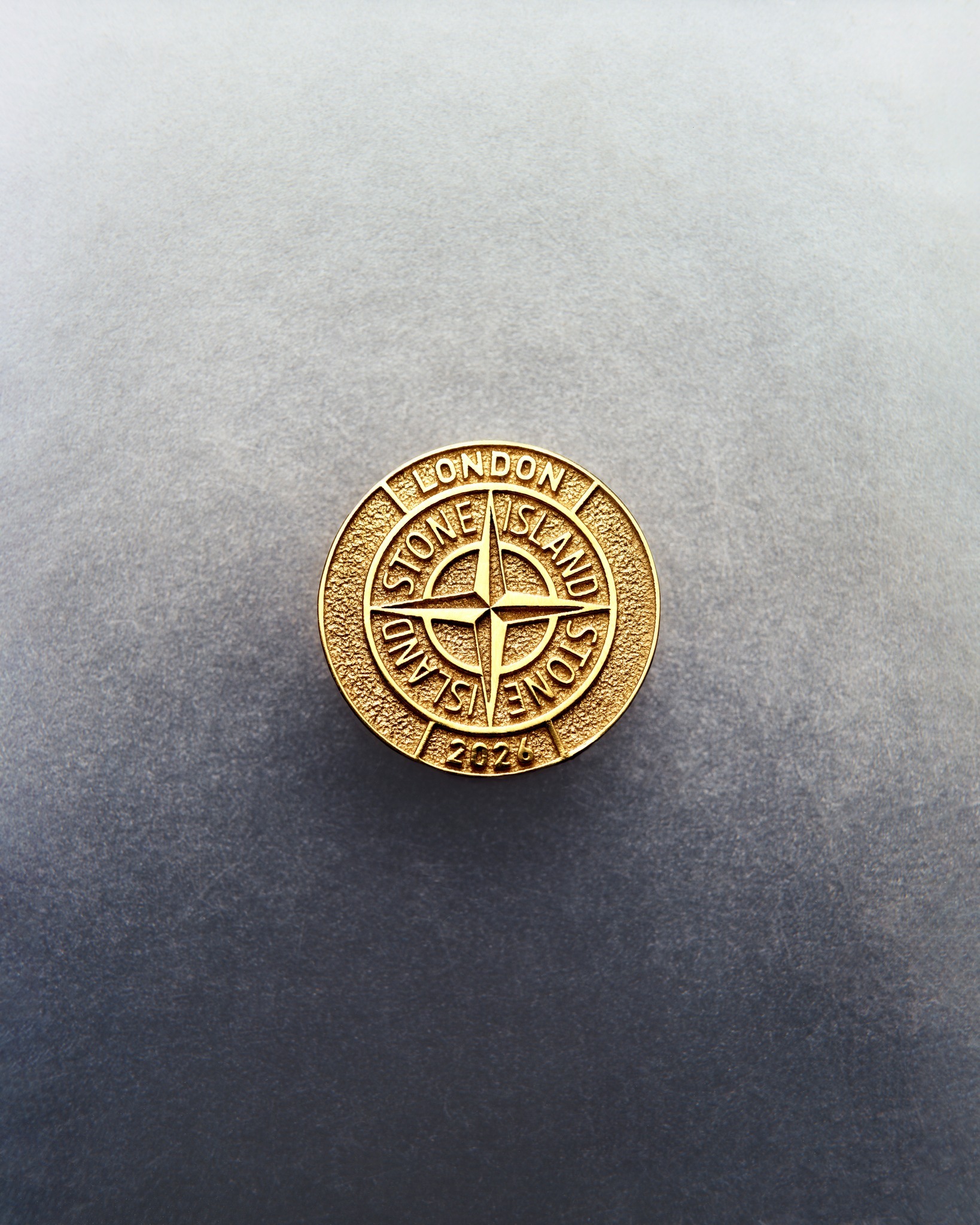 Gold Stone Island round badge with compass logo and 'London' inscription on textured grey background