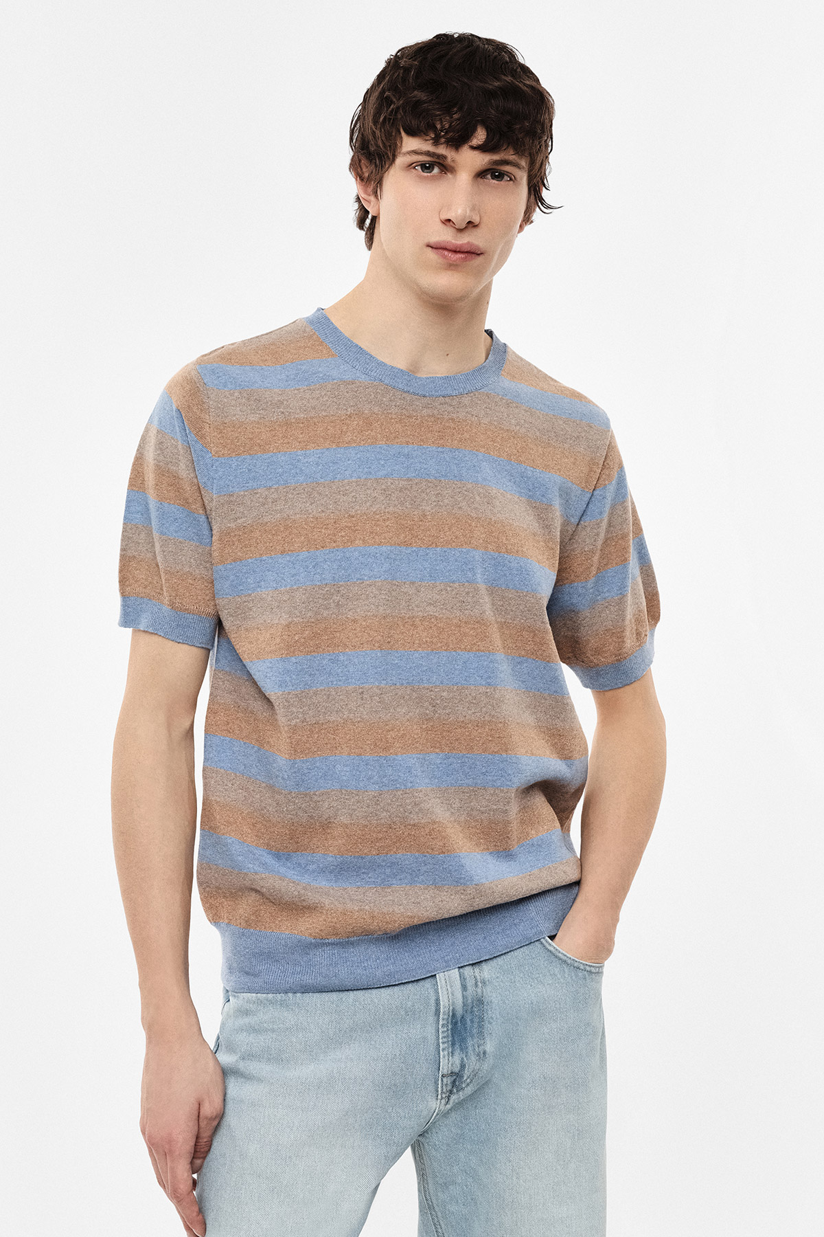 Male model wearing blue and tan striped short-sleeve knit jumper with light-wash jeans, hand in pocket against white background