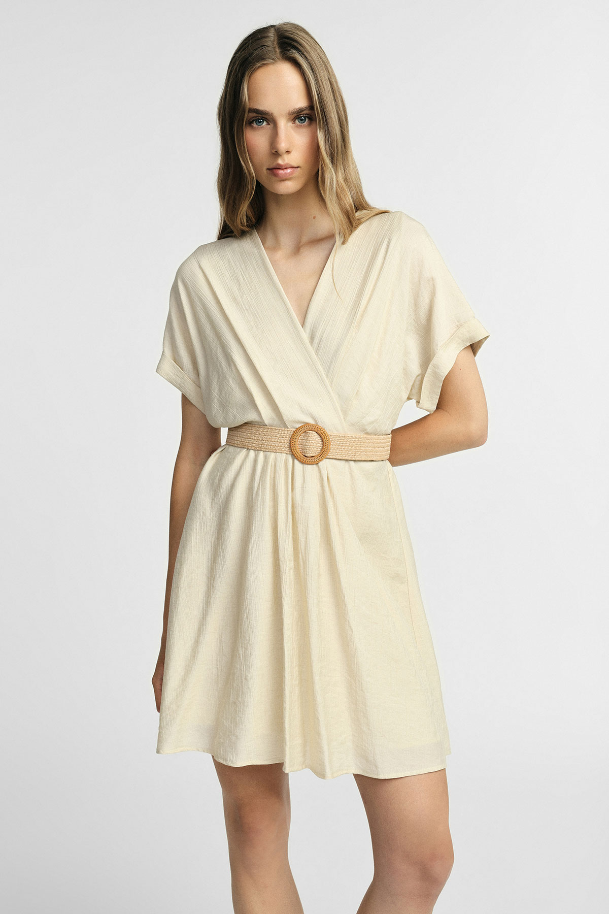 Woman in a cream short wrap dress with short sleeves and a woven belt with round buckle, standing