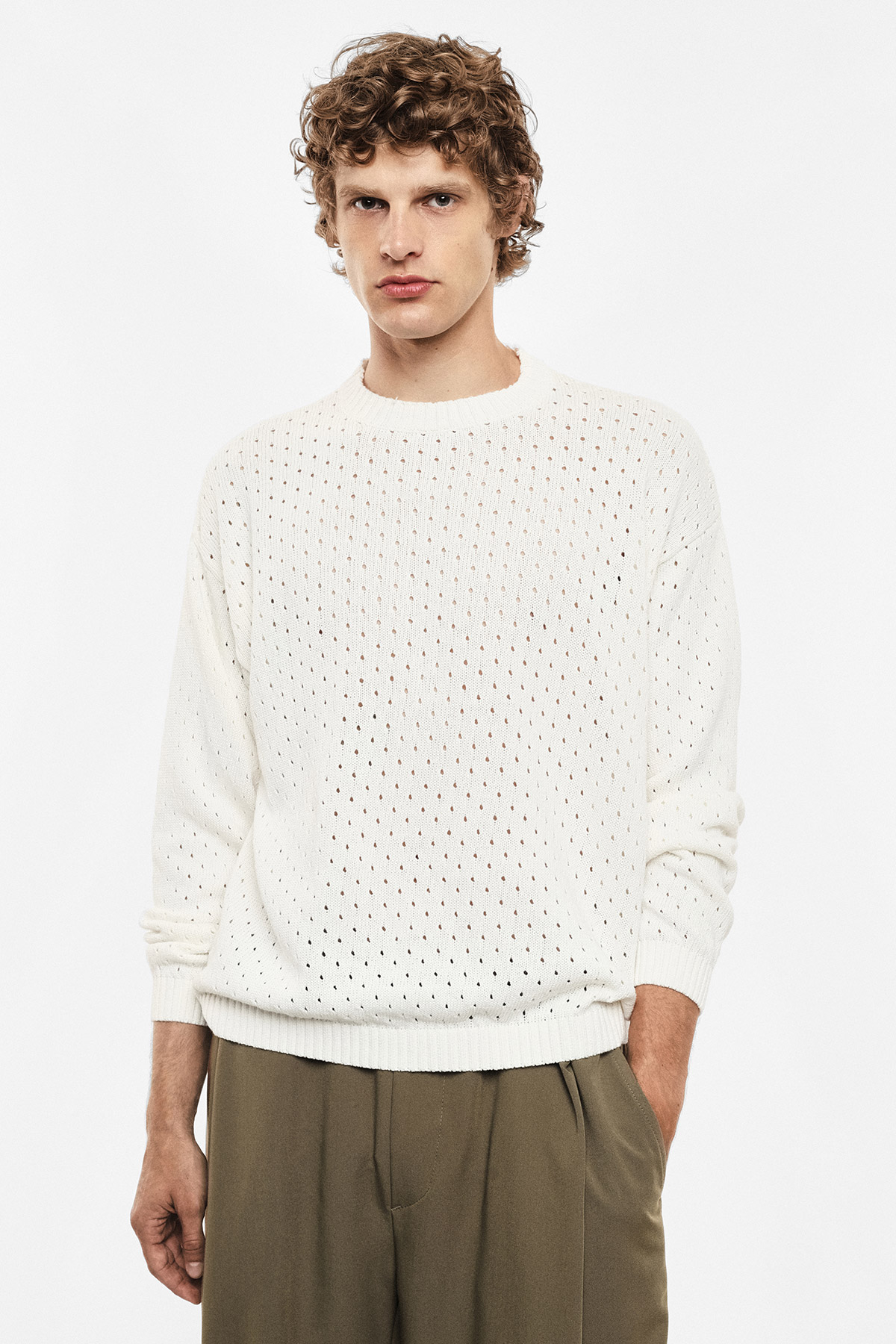 Young man modelling cream open-knit sweater and olive trousers, standing against plain white background, hands in pockets