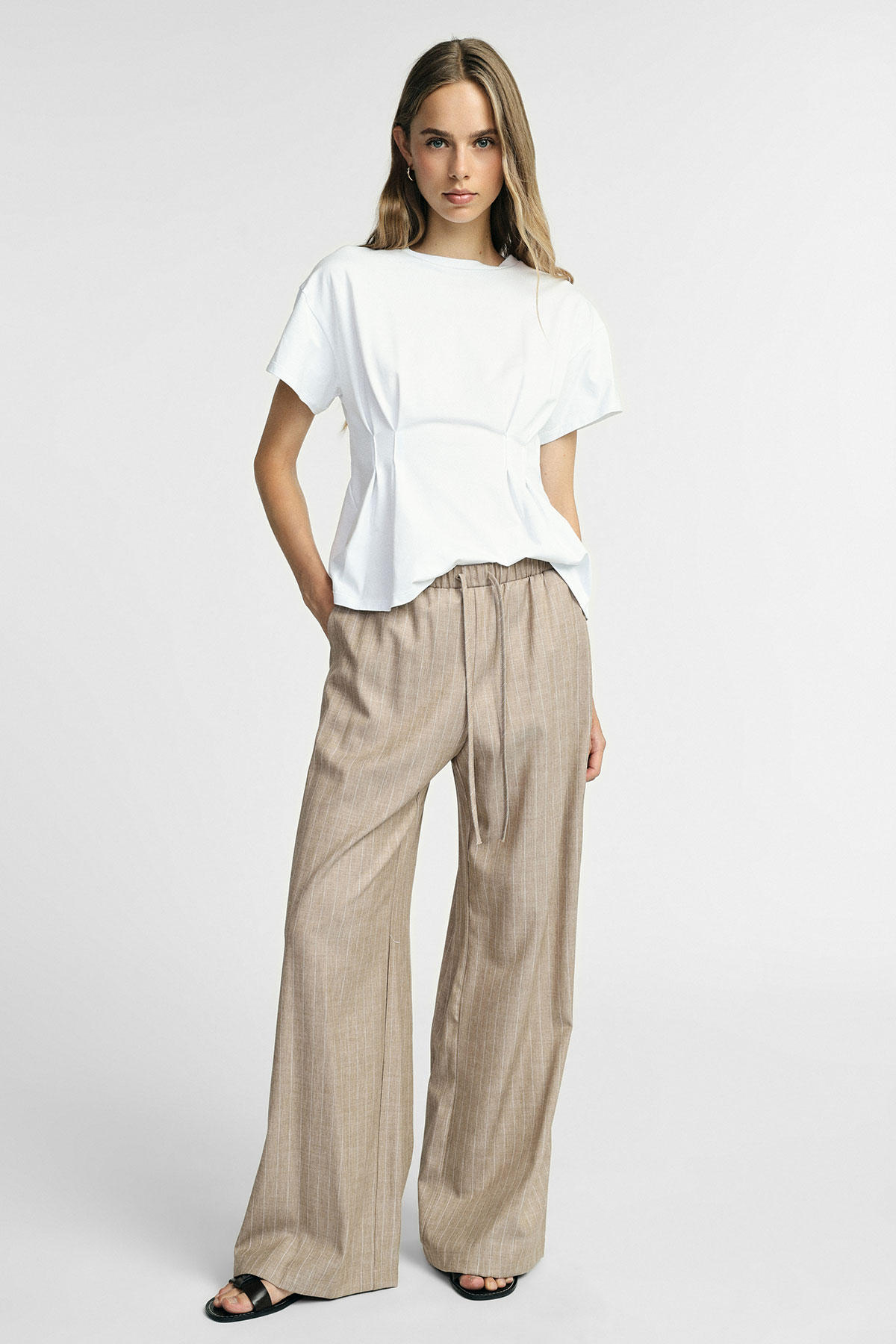 Woman wearing white short-sleeve peplum top and beige wide-leg linen trousers with tie waist, black sandals, studio background