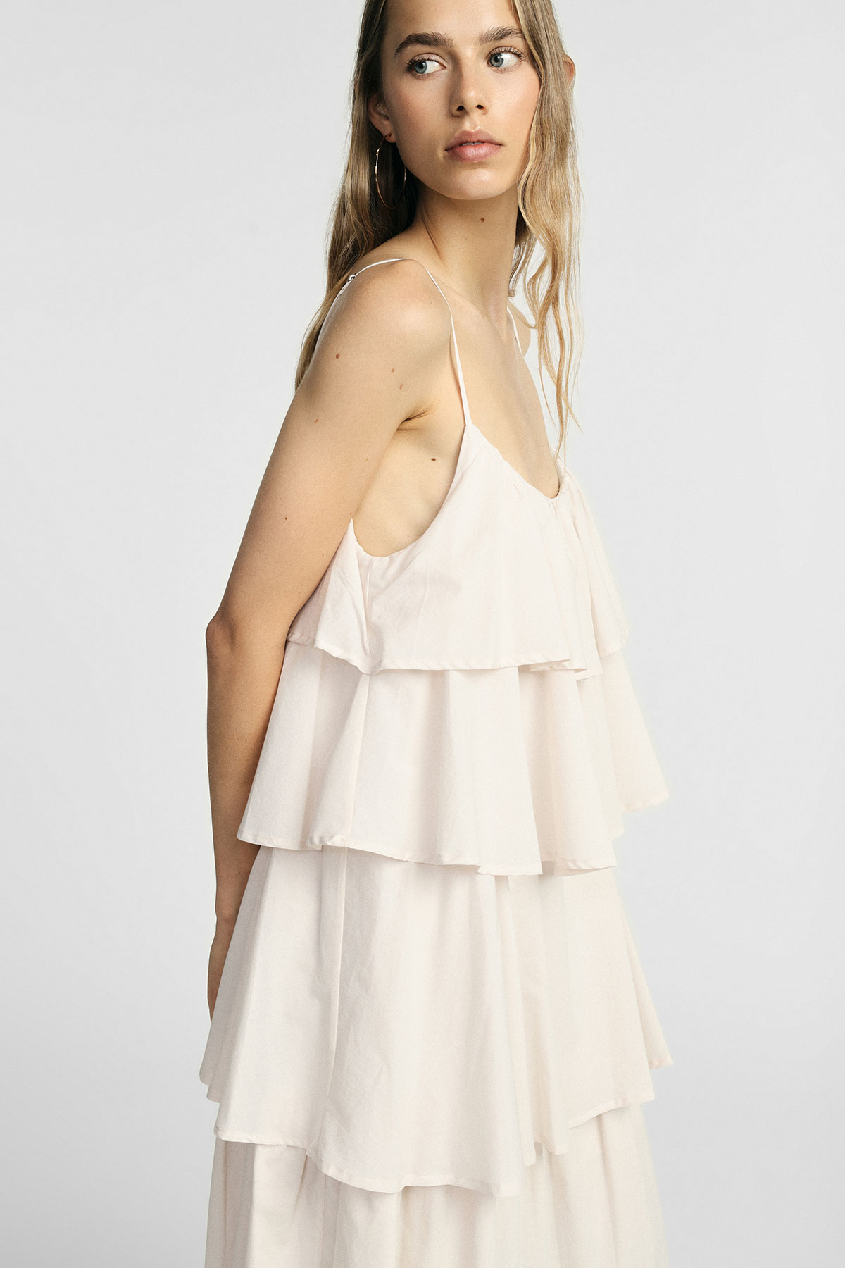 Woman in cream tiered spaghetti-strap summer dress with hoop earring, side pose against a plain pale background