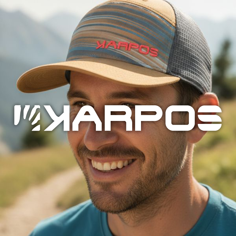 Smiling man wearing a Karpos cap on a sunlit mountain path, close-up portrait with blurred peaks in the background