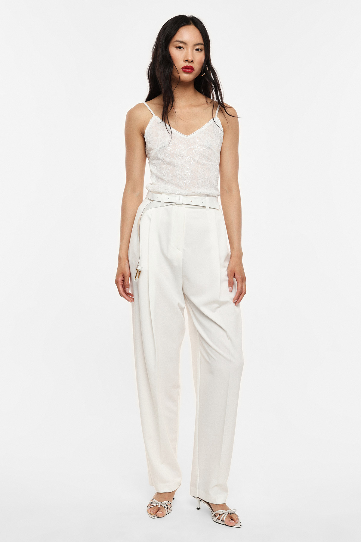 Woman model in white sequin camisole and high-waisted wide-leg white trousers with belt and strappy heels on plain background
