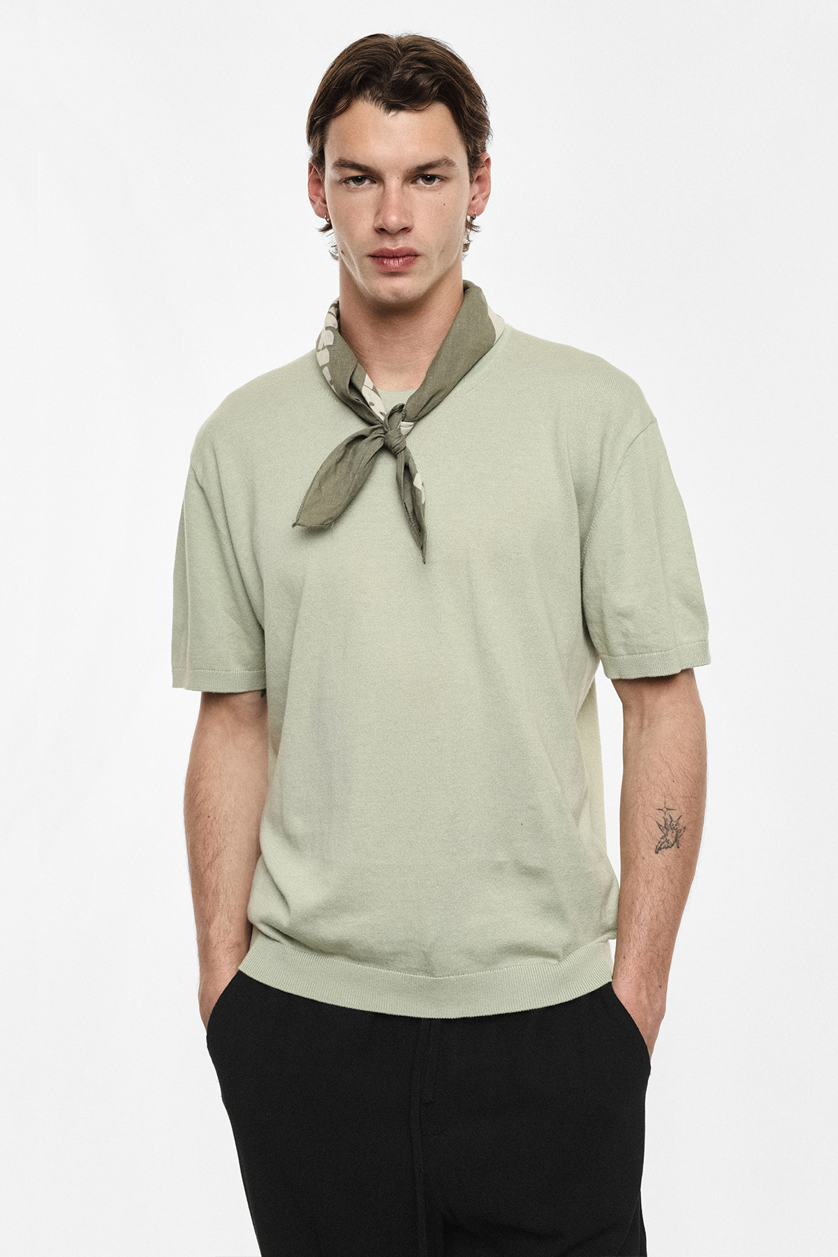 Male model wearing a sage-green short-sleeve knit top with an olive neckerchief and black trousers, hands in pockets