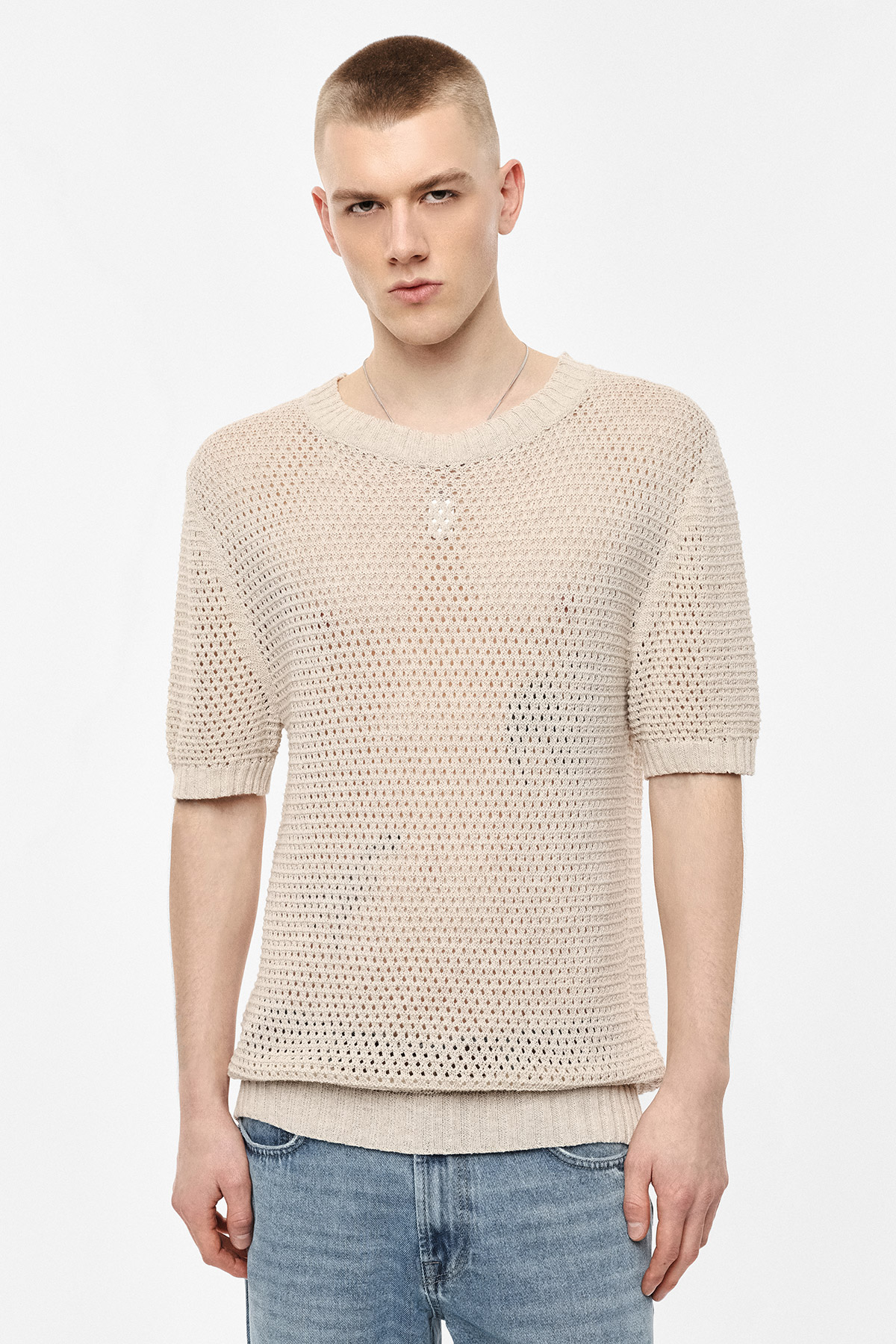 Young man wearing beige open-knit short-sleeve sweater layered over ribbed hem and light blue jeans against white background