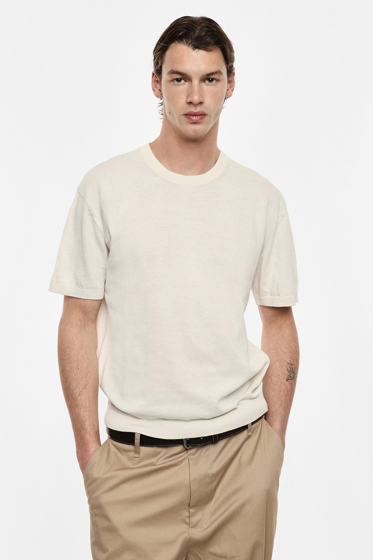 Male model wearing cream short-sleeve knit sweater and khaki trousers, hands in pockets on a white studio background