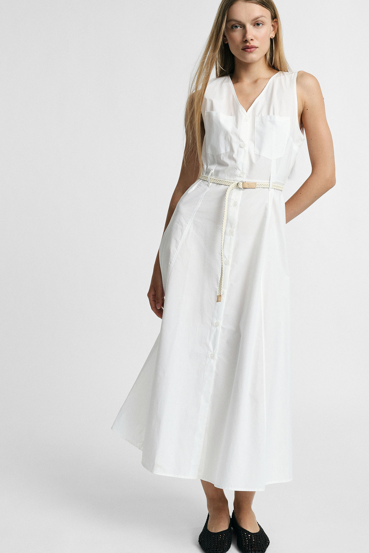 Model wearing sleeveless white button-front midi dress with braided belt and black flats, posed against a plain light background