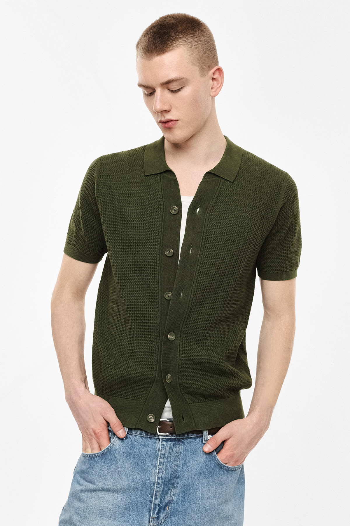 Young man wearing olive green short-sleeve knitted cardigan over white tee and blue jeans, hands in pockets, looking down