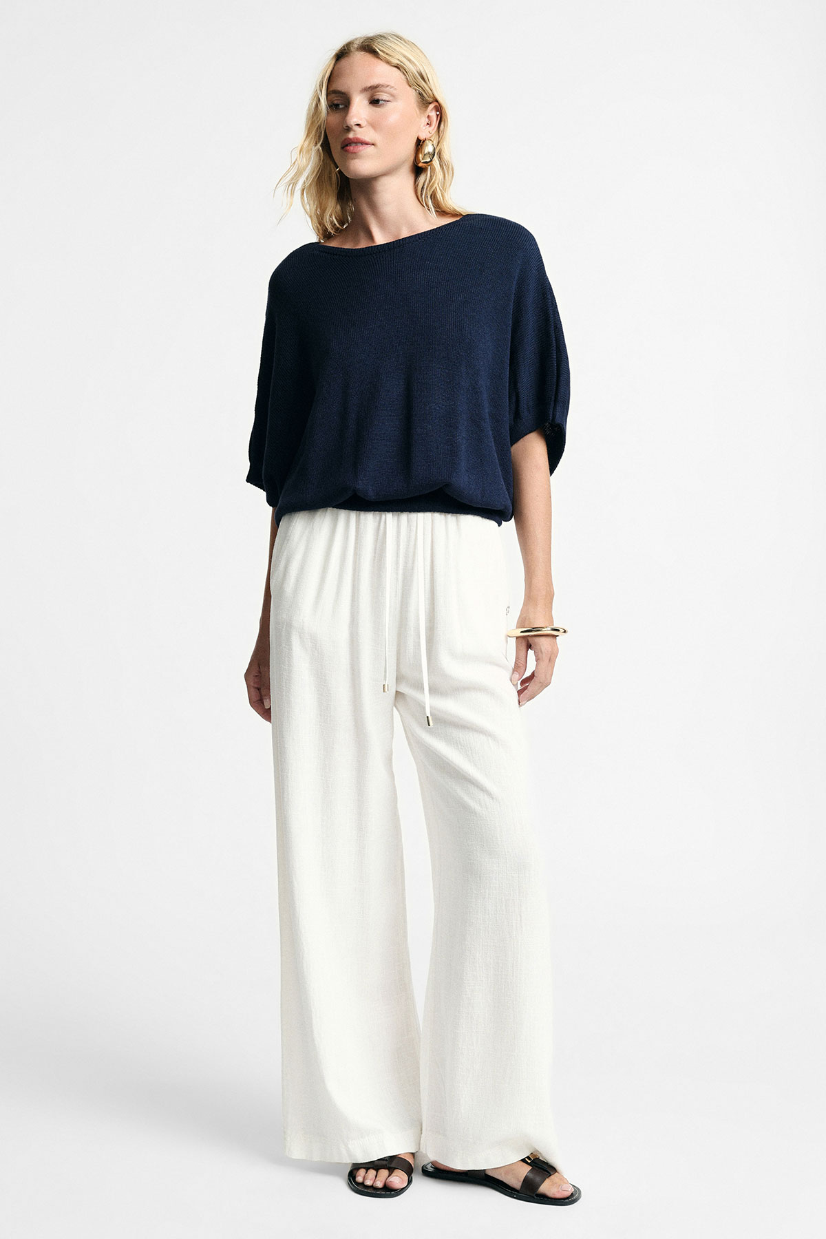 Blonde woman modelling navy loose knit top with short dolman sleeves, white wide-leg drawstring trousers and black sandals on plain background