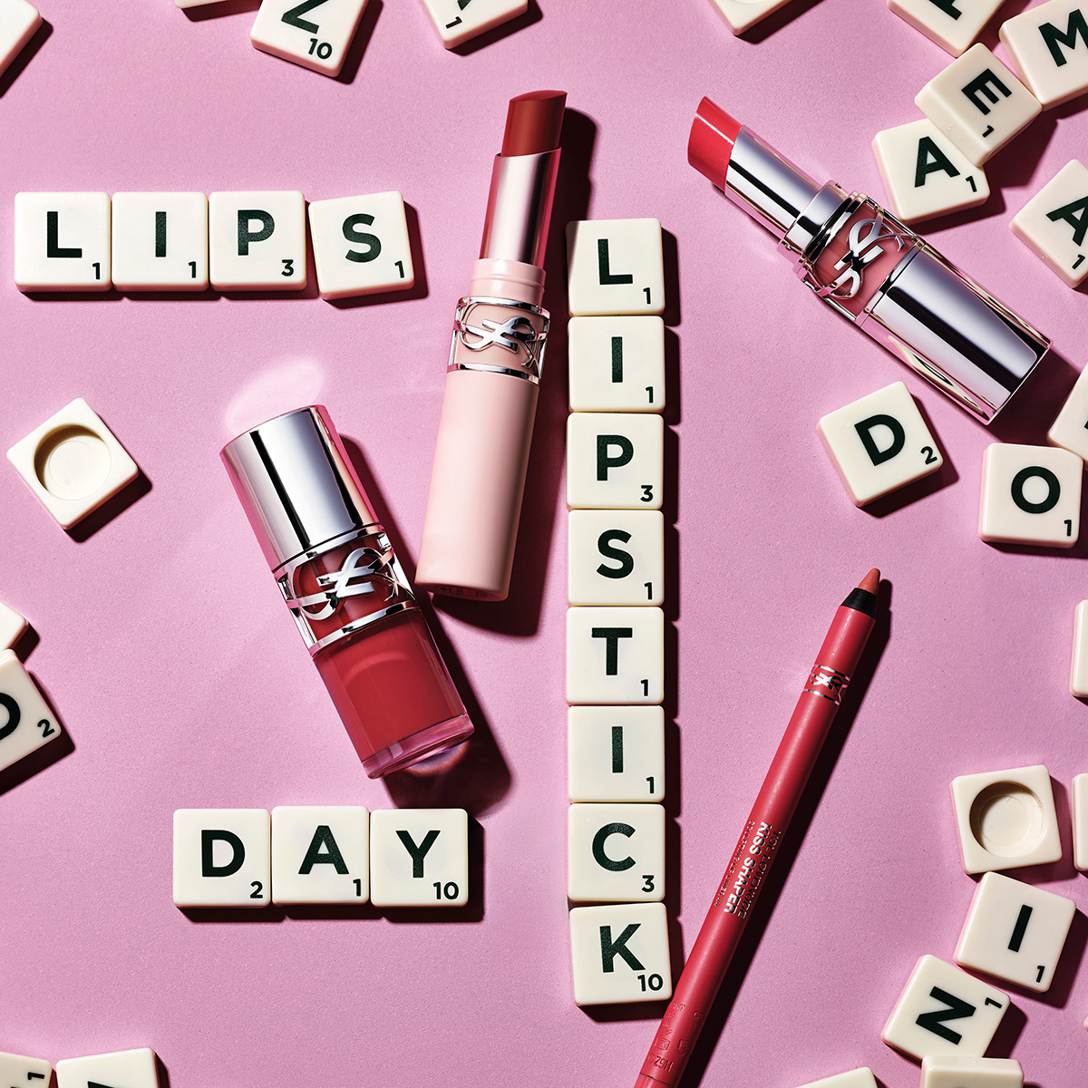 Una selezione di rossetti e una matita labbra YSL disposti su uno sfondo rosa con lettere di Scrabble che compongono "LIPS", "LIPSTICK" e "DAY".