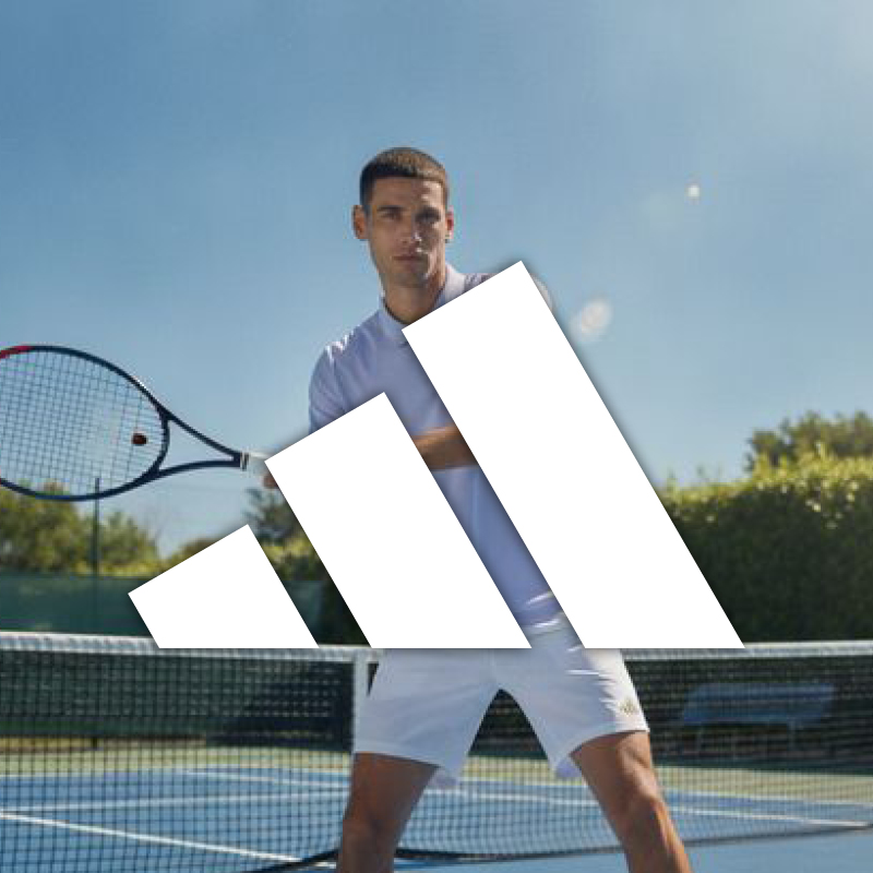 Tennis player in white kit holding a racket on an outdoor court with a large Adidas logo overlay