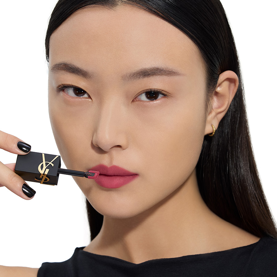 Primo piano di una donna asiatica con smalto nero mentre applica un rossetto liquido opaco color prugna con un applicatore YSL.