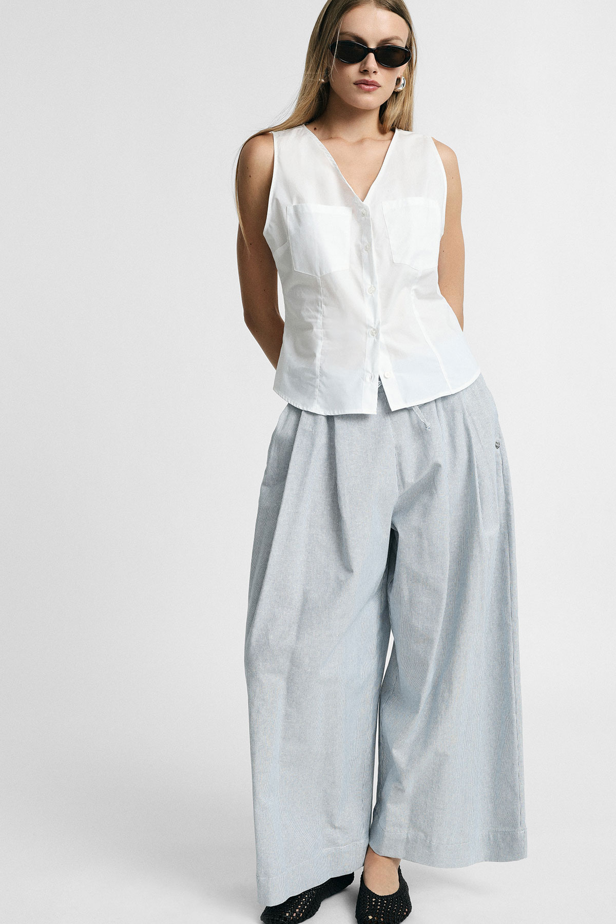 Woman model wearing a white sleeveless buttoned blouse, wide‑leg light blue striped trousers and sunglasses, standing against a plain background