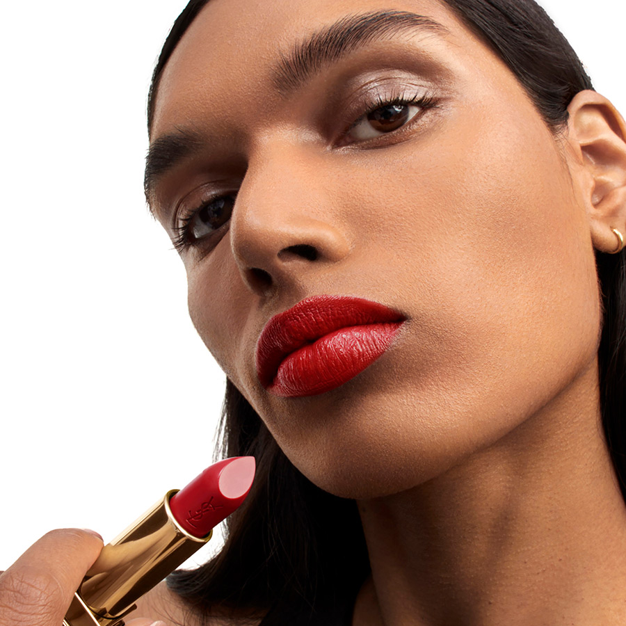 Primo piano di una donna dalla pelle medio-scura che tiene un rossetto rosso intenso YSL vicino alle sue labbra perfettamente truccate.
