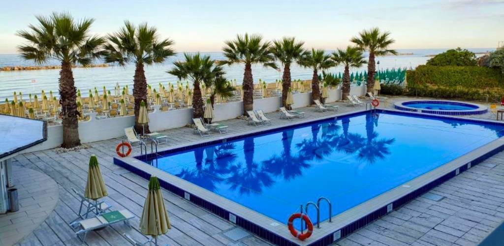 Outdoor hotel swimming pool with palm trees, sunbeds and closed parasols on a wooden deck overlooking calm sea and beach.