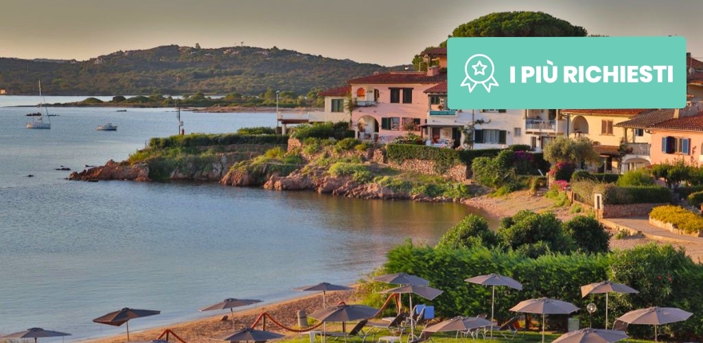 Calm Mediterranean bay with sailboats, rocky cove and seaside villas above a sandy beach lined with loungers and parasols