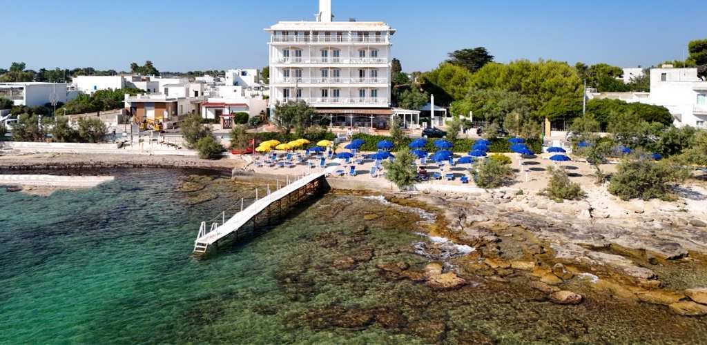Seafront hotel with wooden pier, rocky shoreline and sandy beach lined with blue and yellow umbrellas and turquoise Mediterranean water