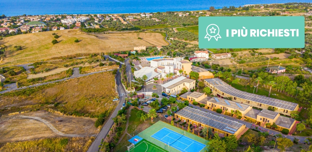 Aerial view of coastal Mediterranean resort with tennis courts, solar-panelled buildings, surrounding fields and town by the sea