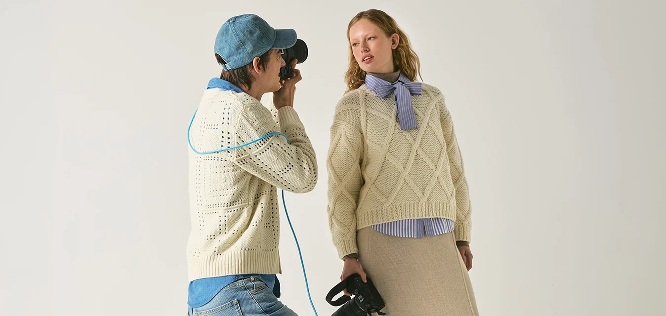 Fotografo con cappellino e maglione scatta una foto a una donna con maglione di lana e gonna beige che tiene degli stivali neri su sfondo uniforme.