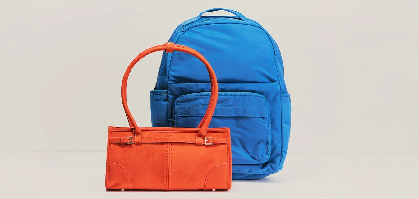 Zaino blu posizionato dietro a una piccola borsa arancione con due manici su sfondo beige chiaro.