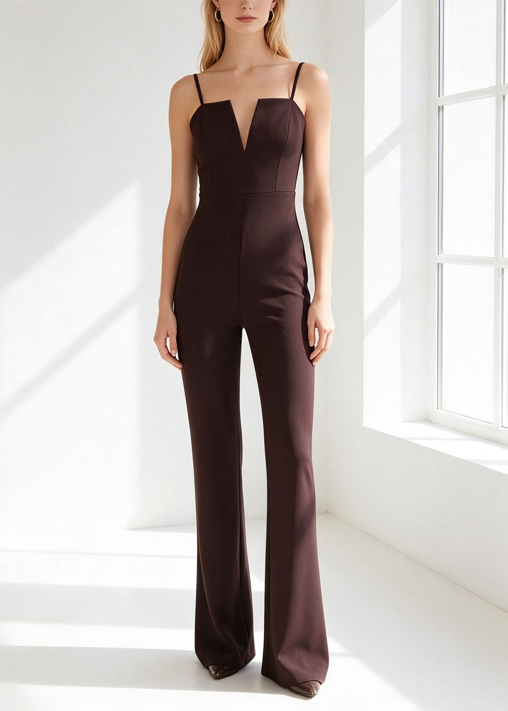 Woman in a chocolate-brown fitted jumpsuit with thin straps, deep V neckline and flared legs, standing by a sunlit window
