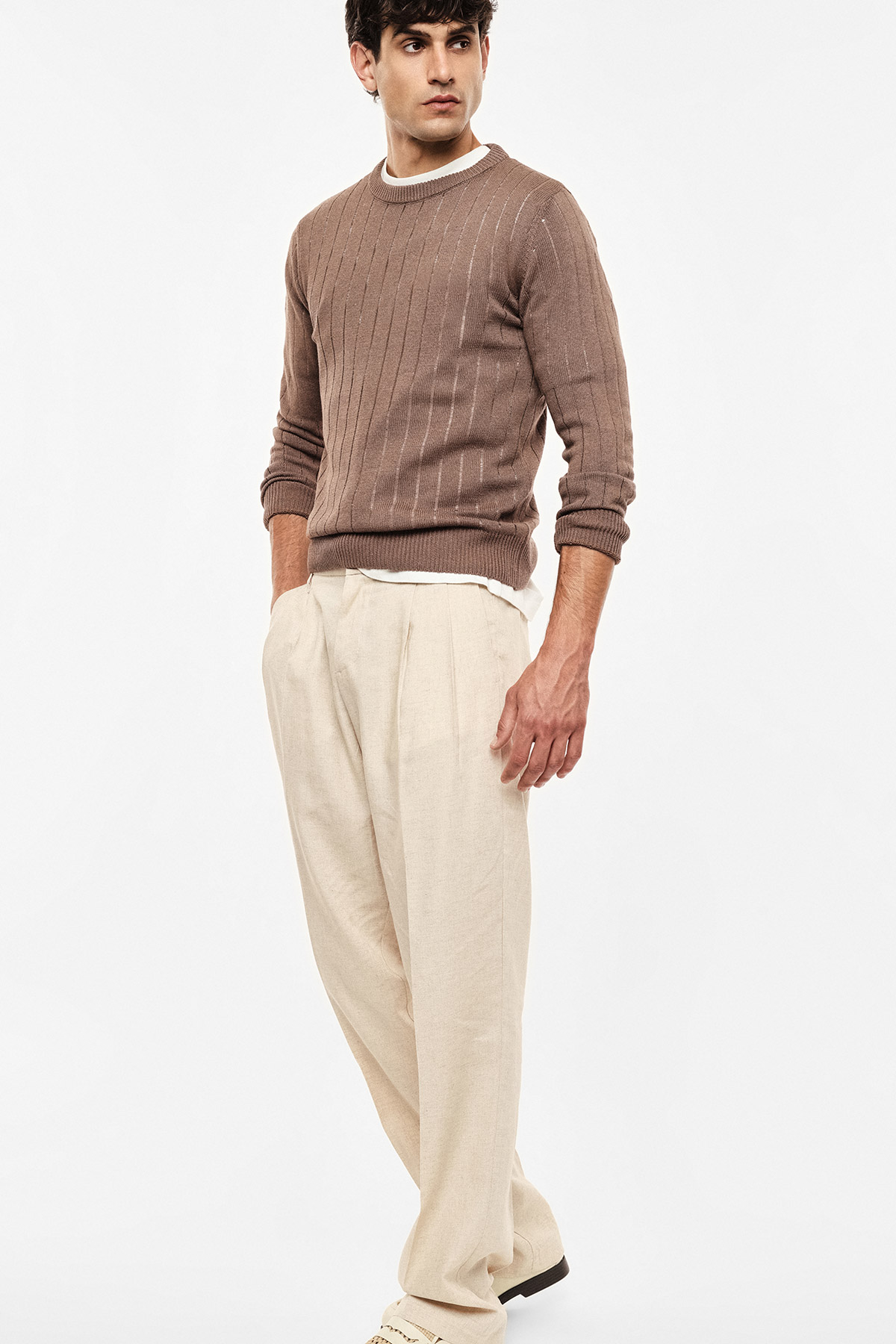 Male model wearing a brown cable-knit jumper and beige pleated trousers against a white background, casual menswear look