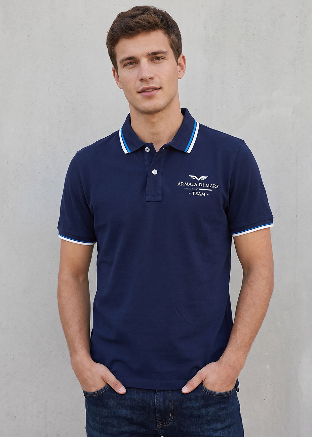 Young man in navy Armata di Mare polo with white-blue striped collar and cuffs, hands in jeans pockets against concrete wall