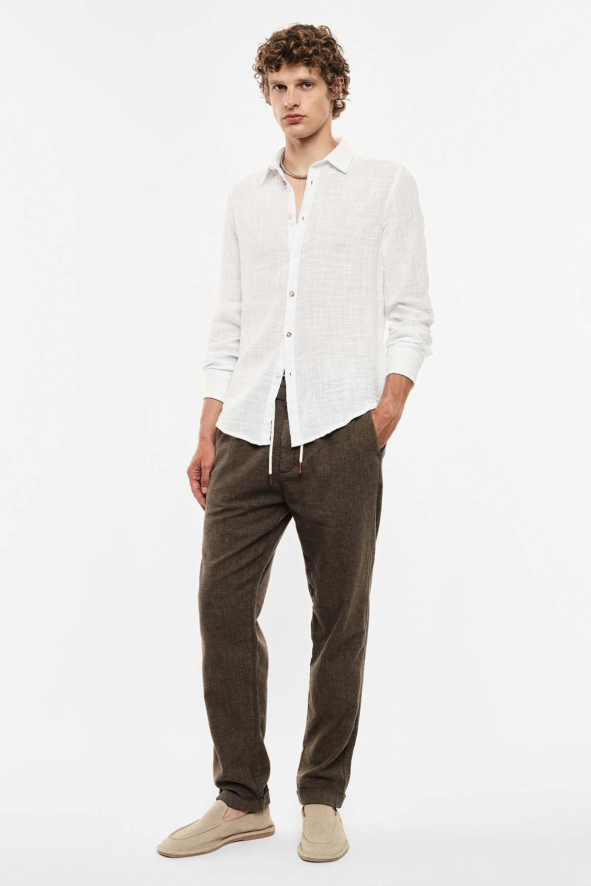 Young man in white linen shirt, brown drawstring casual trousers and beige slip-on shoes on white background