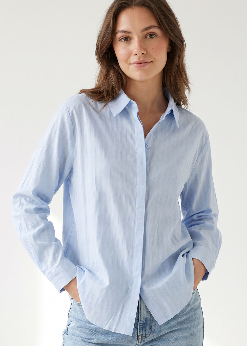 Woman modelling light blue button-up shirt and jeans with hands in pockets against neutral background