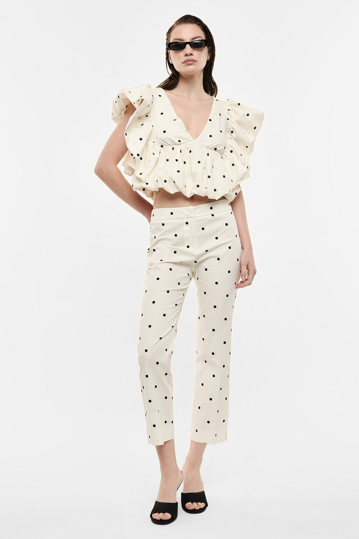 Model wearing cream polka-dot co-ord: ruffled V-neck crop top and matching high-waist trousers, black sunglasses and heels