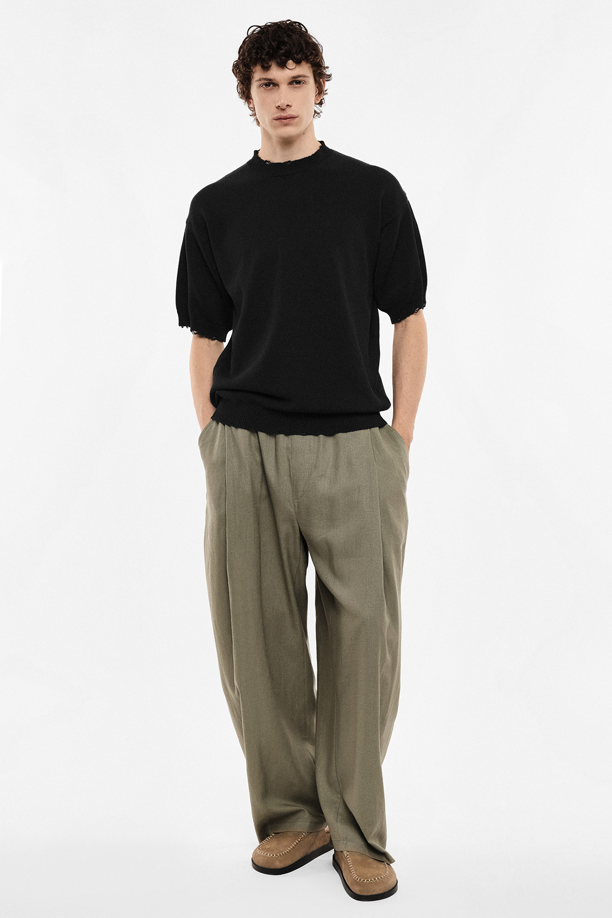 Full-length studio portrait of a man wearing a black short-sleeve knit, olive wide-leg trousers and tan loafers, hands in pockets