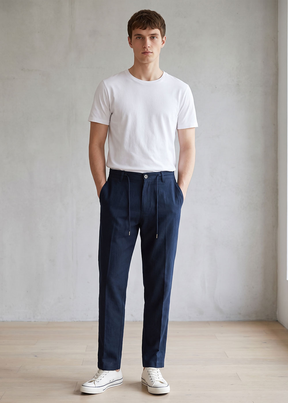 Young man modelling a casual outfit: white crew-neck T-shirt, tailored navy trousers with drawstring and white trainers against a plain studio wall