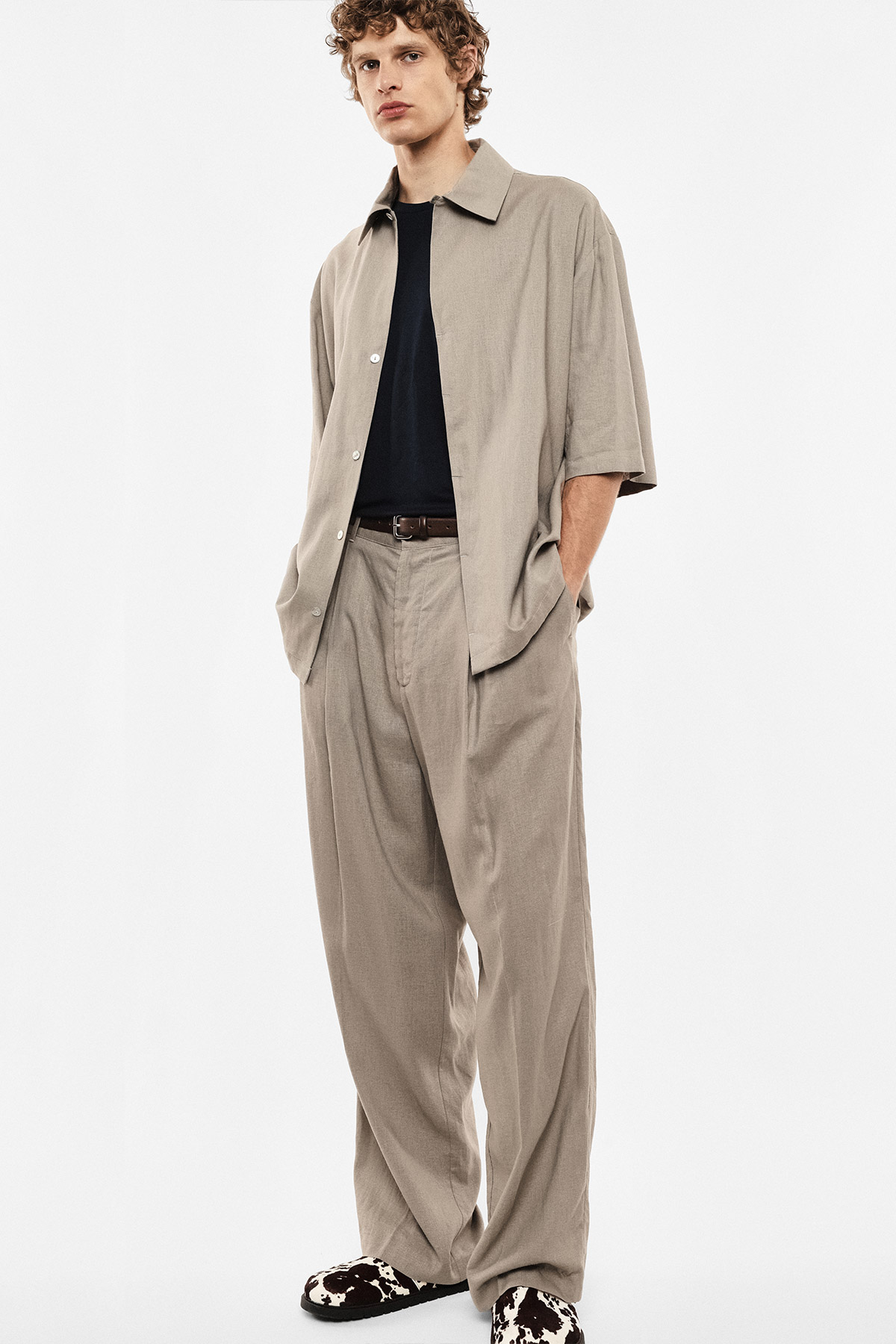 Male model in beige relaxed-fit short-sleeve shirt and wide trousers, black T-shirt, patterned slip-on shoes, hands in pockets