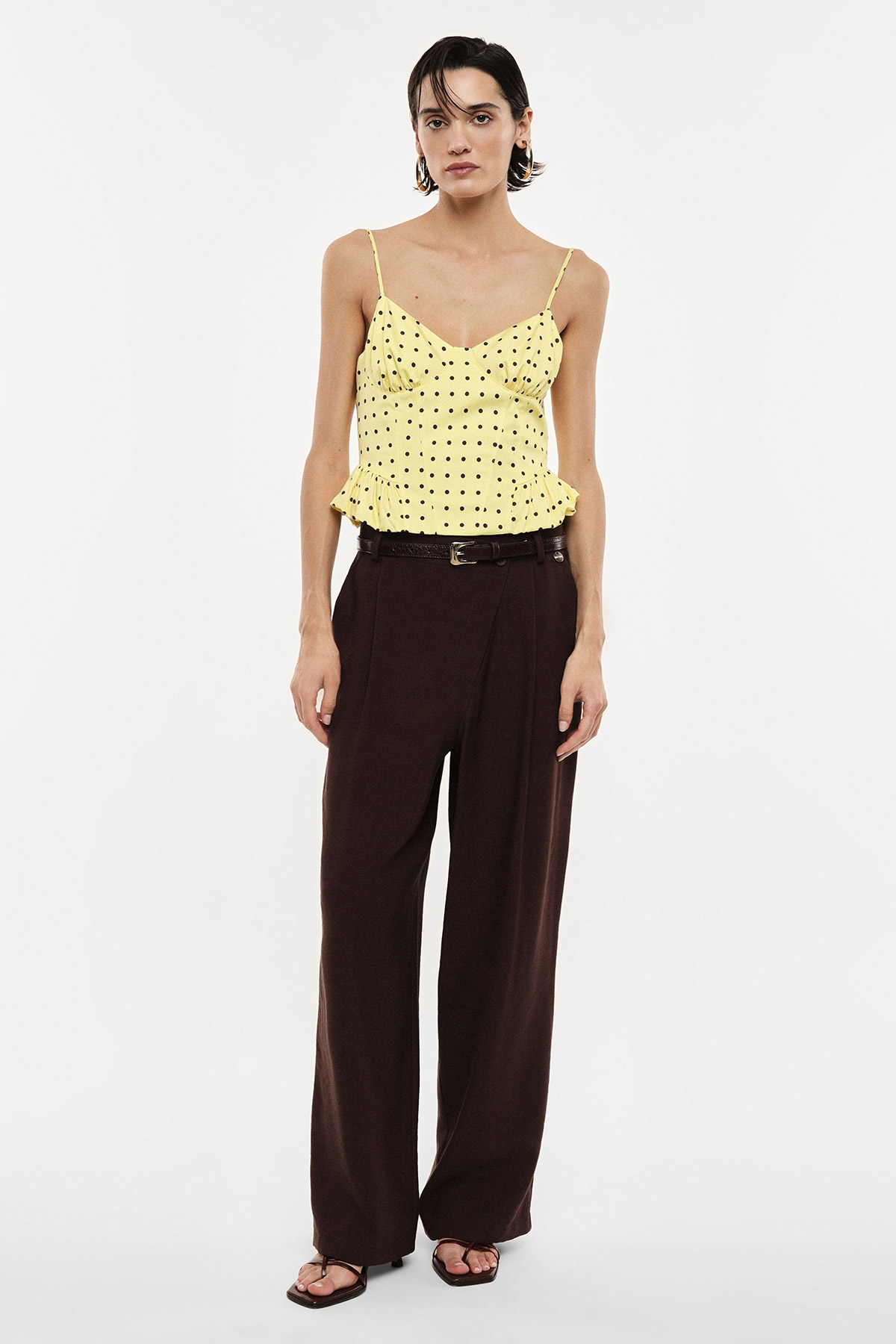 Full-length shot of woman modelling yellow polka-dot ruffled camisole and wide-leg brown trousers on white background