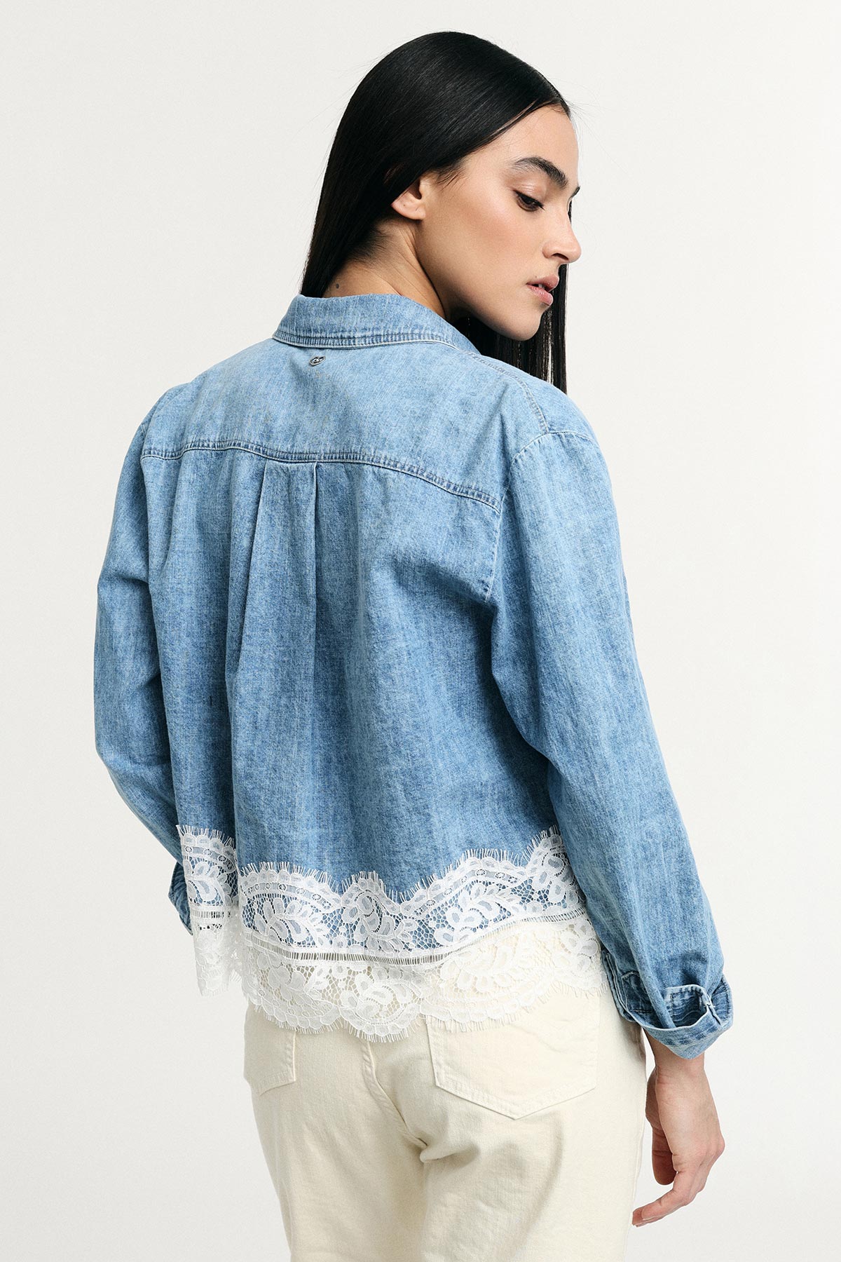 Back view of woman wearing cropped light denim shirt with white lace hem and rolled cuffs, cream trousers