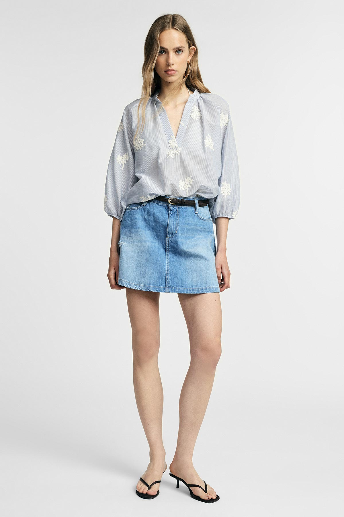 Woman in light-blue embroidered blouse tucked into a denim mini skirt with black belt and strappy sandals