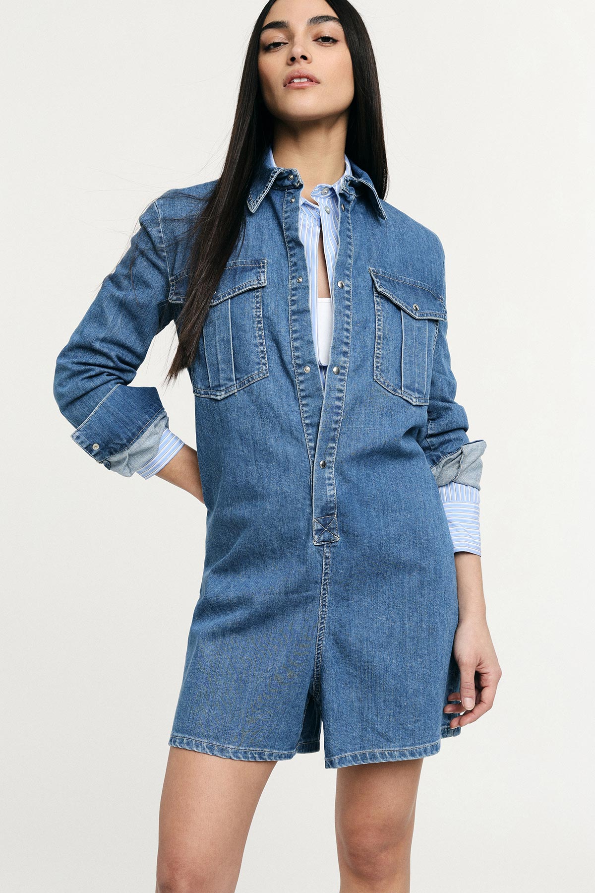 Woman modelling blue denim shirt-dress layered over striped shirt, one hand on hip against plain light background