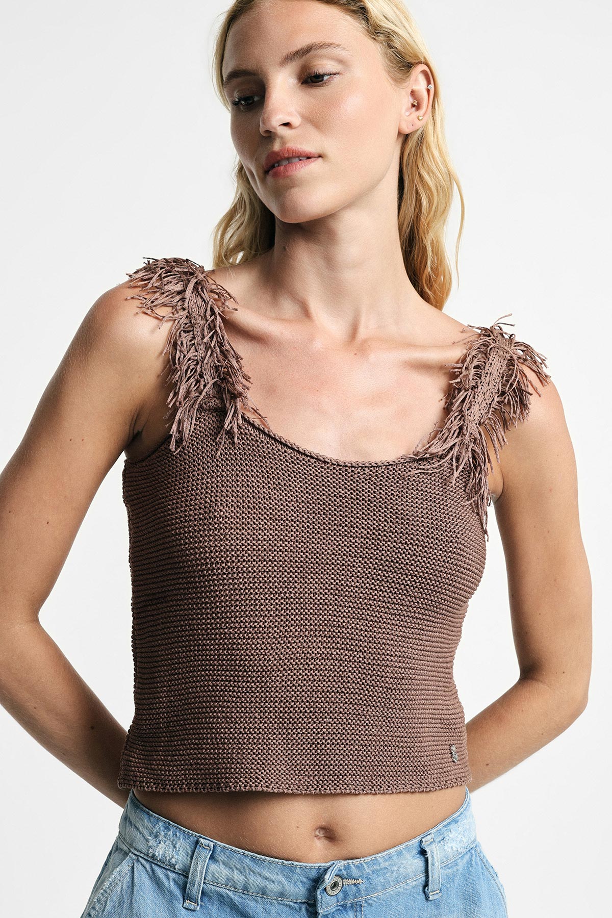 Blonde woman wearing a brown knitted cropped tank top with fringe shoulder straps paired with light blue jeans, front view