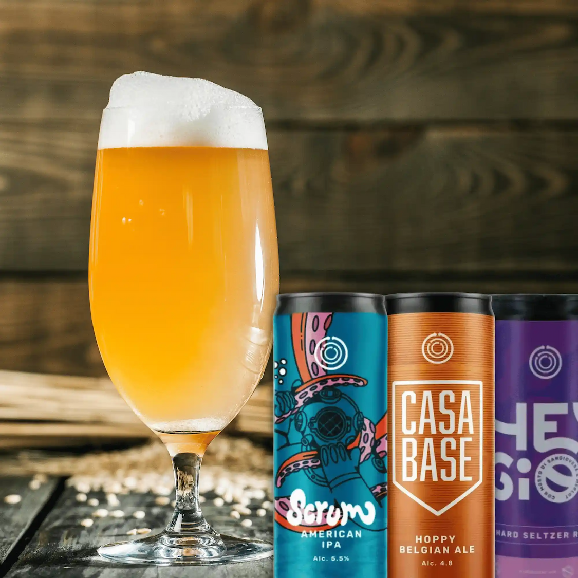 A glass of beer next to three cans of craft beer: Scrum American IPA, Casa Base Hoppy Belgian Ale, and Hey Gio Hard Seltzer.