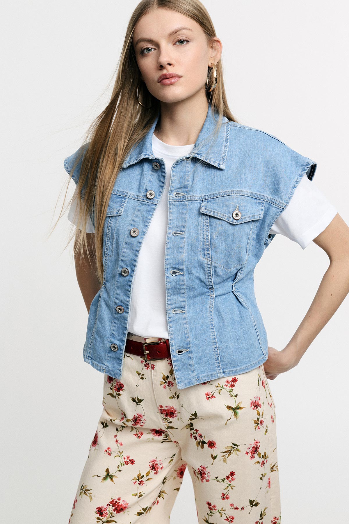 Woman wearing light denim sleeveless jacket over white T-shirt and floral trousers, hand on hip, studio portrait